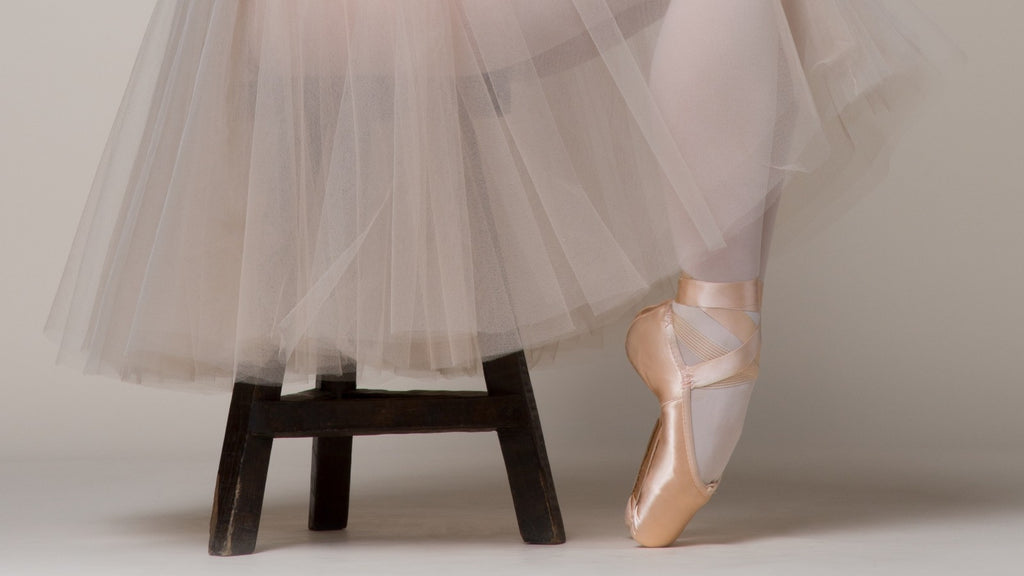 A ballet dancer sitting on a stool with feet in pointe position wearing pointe shoes and a tutu