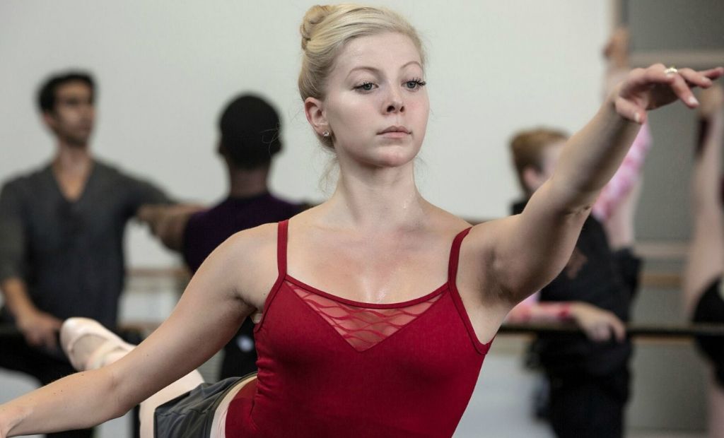 A female ballet dancer wearing a red camisole leotard practising ballet at the barre in the classroom with help from her teacher